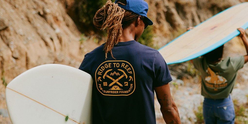 A surfer walks carrying a mid-length board, wearing a navy blue Surfrider and Protect Our Winters collab shirt that reads "Ridge to Reef."