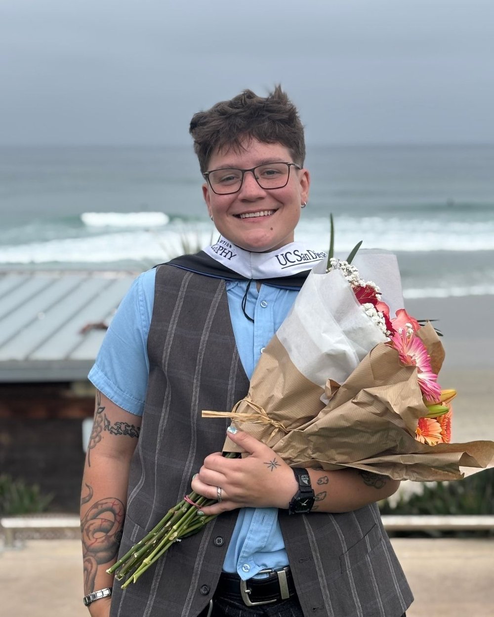 Addie poses at the beach during their UC San Diego graduation, holding a bouquet of flowers.