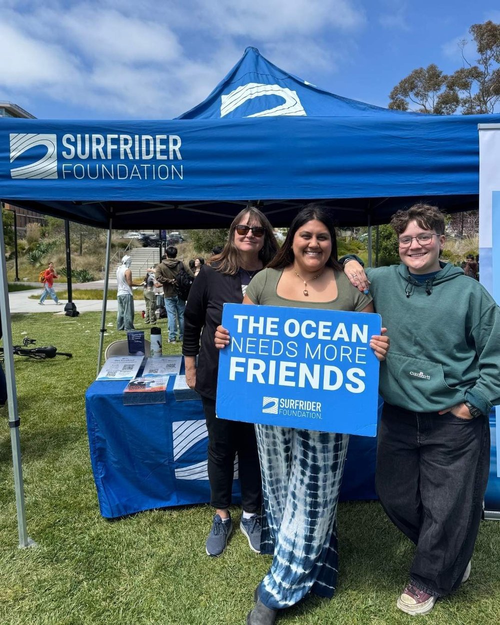 Addie poses under a blue Surfrider tent. Joana Guerra, chapter manager, holds a sign that reads "the ocean needs more friends."