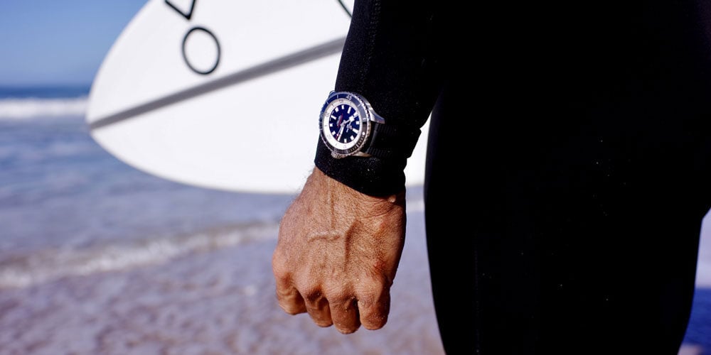 A person holding a surfboard, wearing a Brietling watch, looking at the ocean