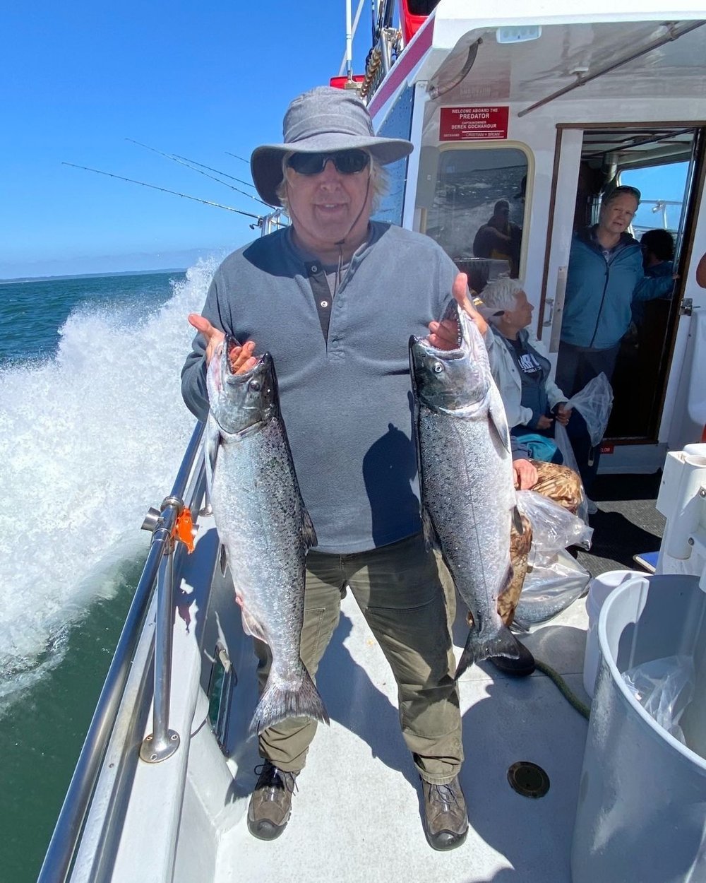 Bruce on a boat holding two large salmon. He is near the rail with the ocean on his right side.