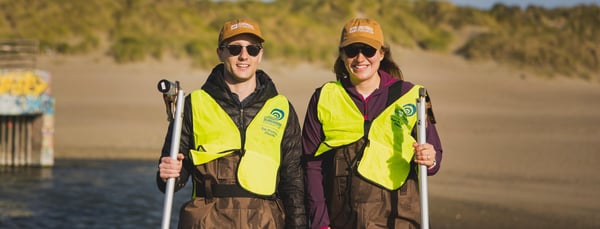 Kyle on left, Amanda on right, pose in hi-viz vests and waders as they prepare to test ocean water quality. They are wearing light brown 