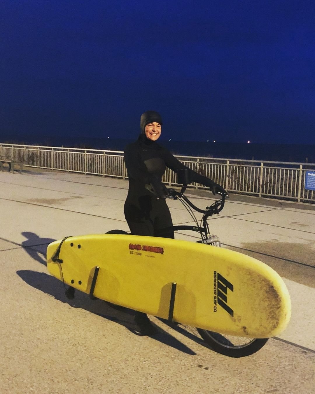 Corinne wearing a wetsuit, on a bicycle, with a surfboard rack. It's still dark, so she's on her way to a dawn patrol session.