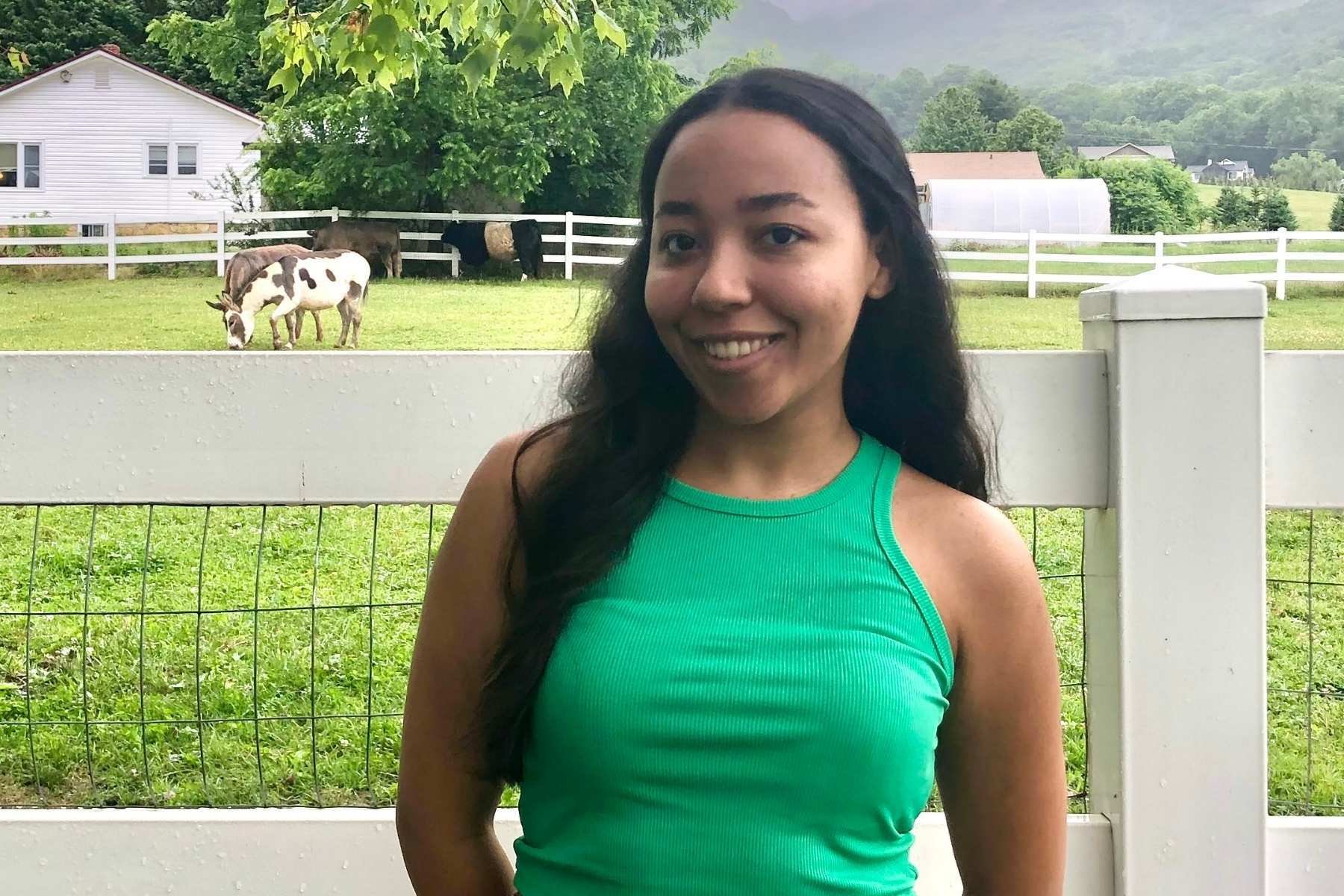 Elianny is wearing a green tank top standing in front of a white fence. In the background there are some farm animals grazing.