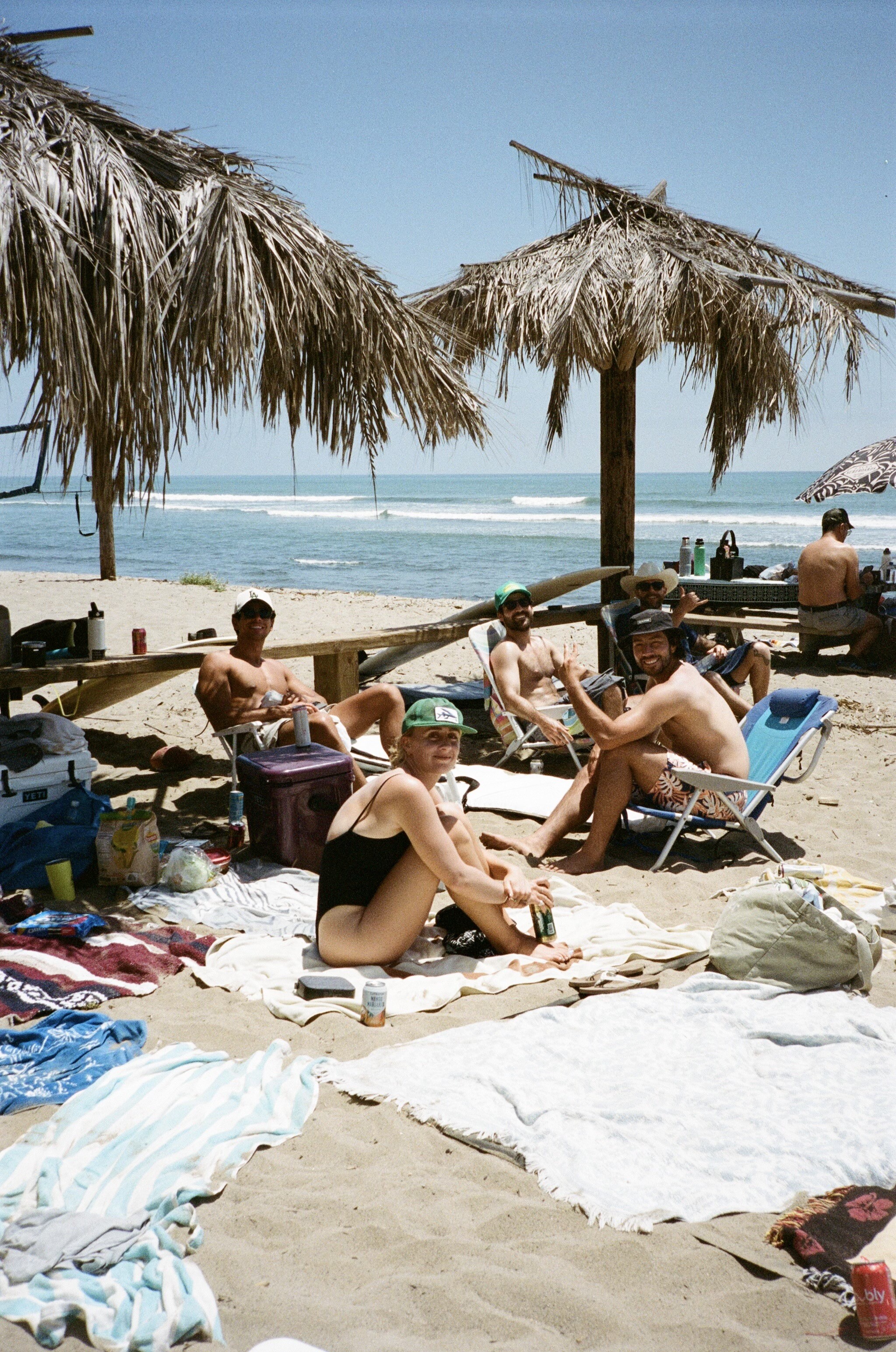 beach towels cover the sand where friends are seated, smiling, with palm frond covered umbrellas and the sparkeling blue ocean behind
