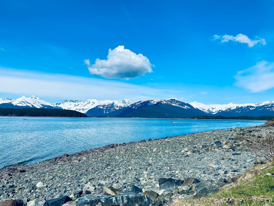 A bright blue day on a rocky beach and bright blue glacial waters with snow capped mountains in the background - photo credit Kathrine Coonjohn from Unsplash 
