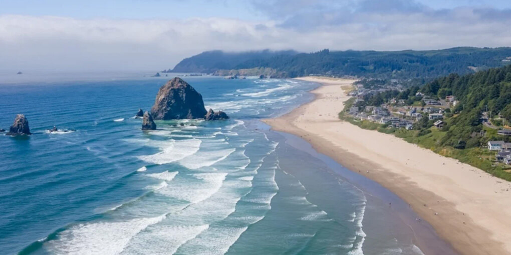 An aerial photograph of Cannon Beach, Oregon, waves peeling, a slightly cloudy sky, and a green coastline.