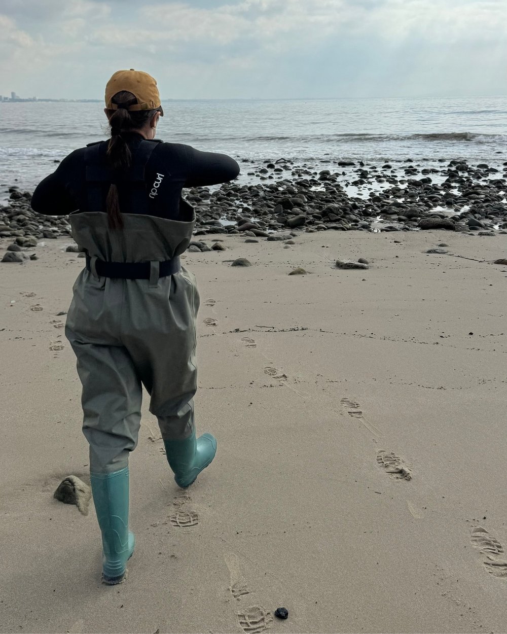 A woman approaches the water's edge in waders preparing to take a water sample.