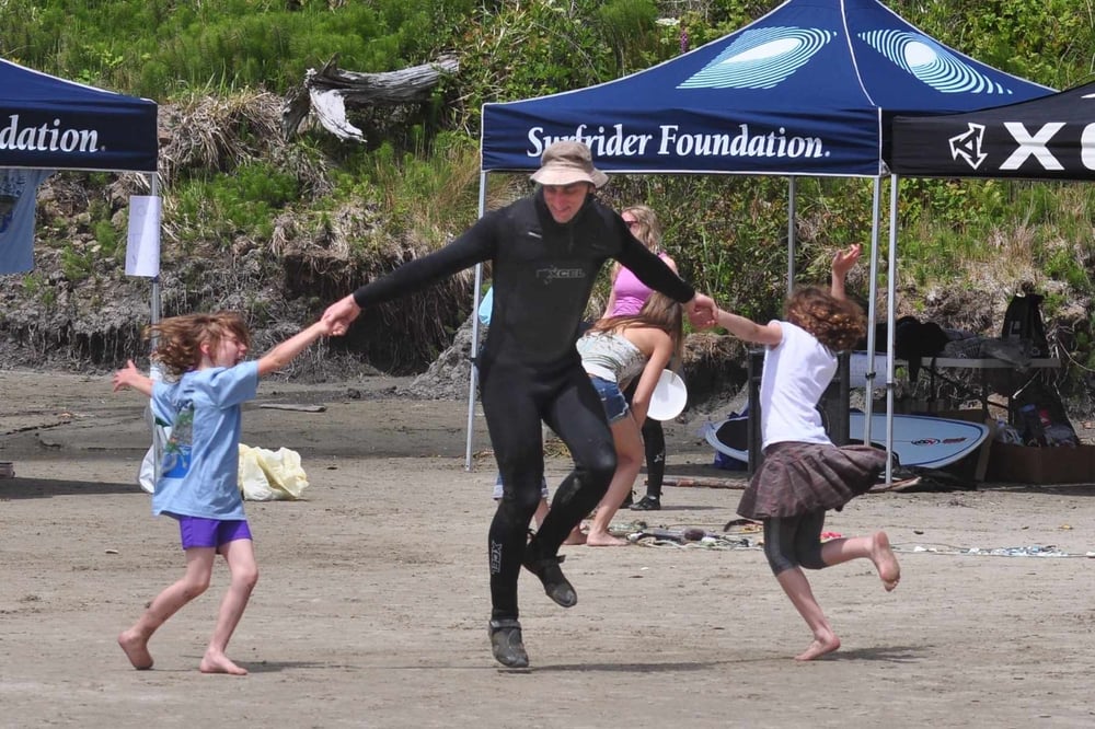 Wearing a wetsuit, Vince dances with volunteers in front of a Surfrider tent.