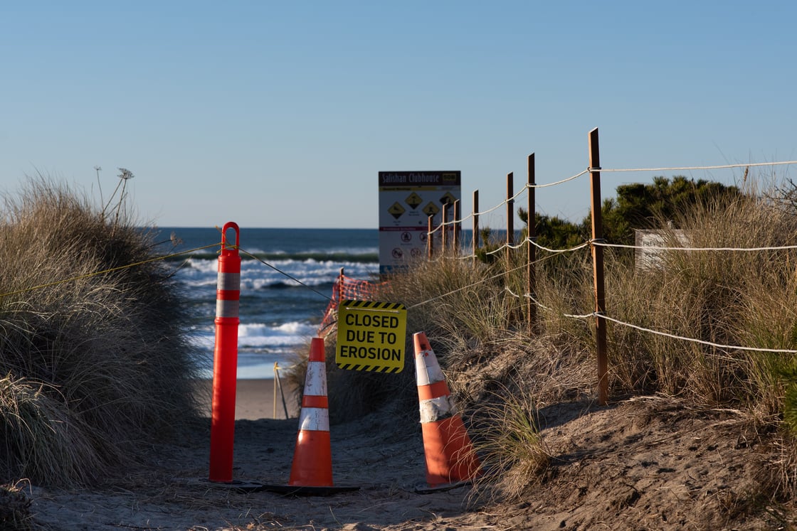 a beach access trail is blocked by cones and a sign reading "closed due to erosion"