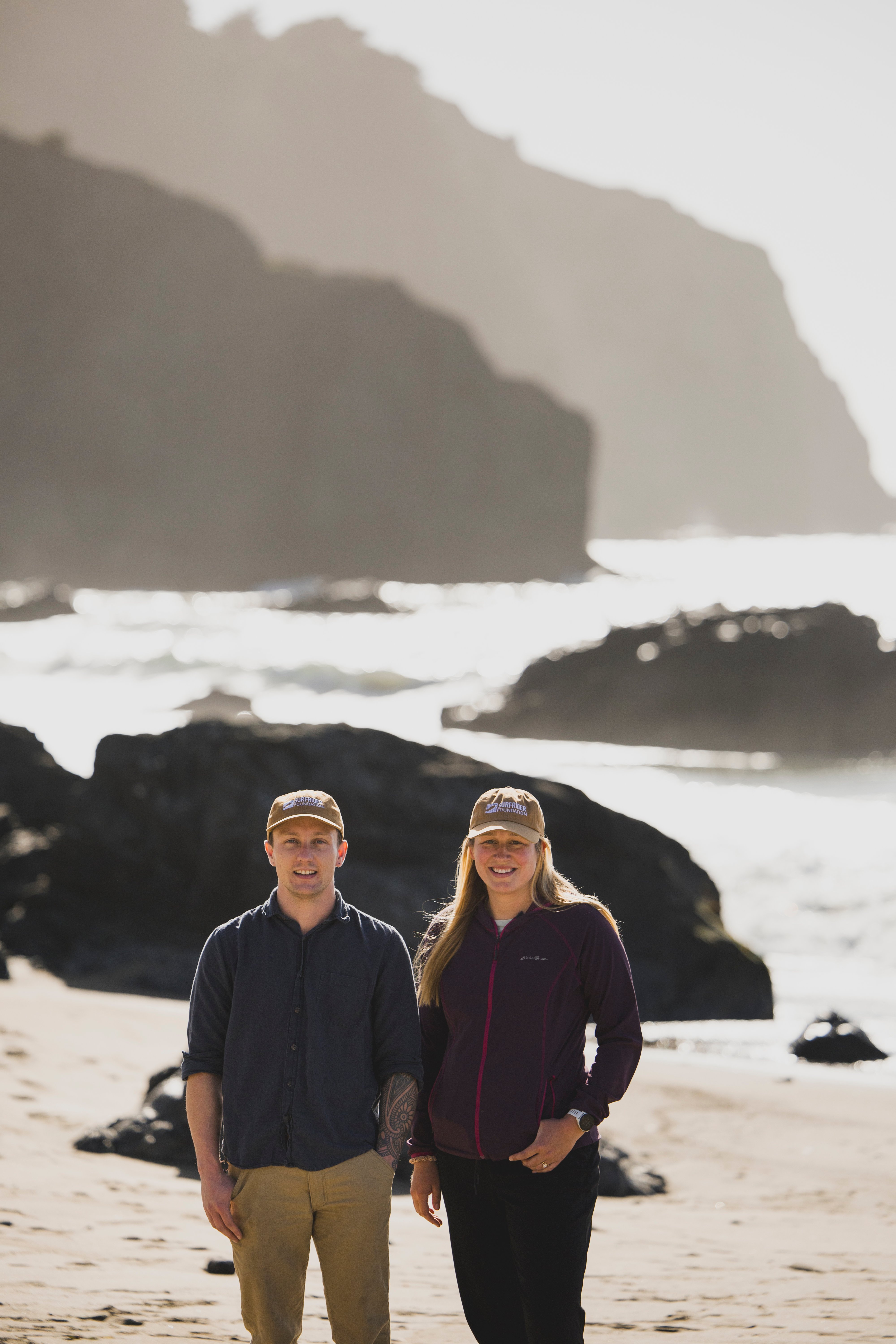Kyle and Amanda pose side by side at the beach with rocky cliffs in the background.