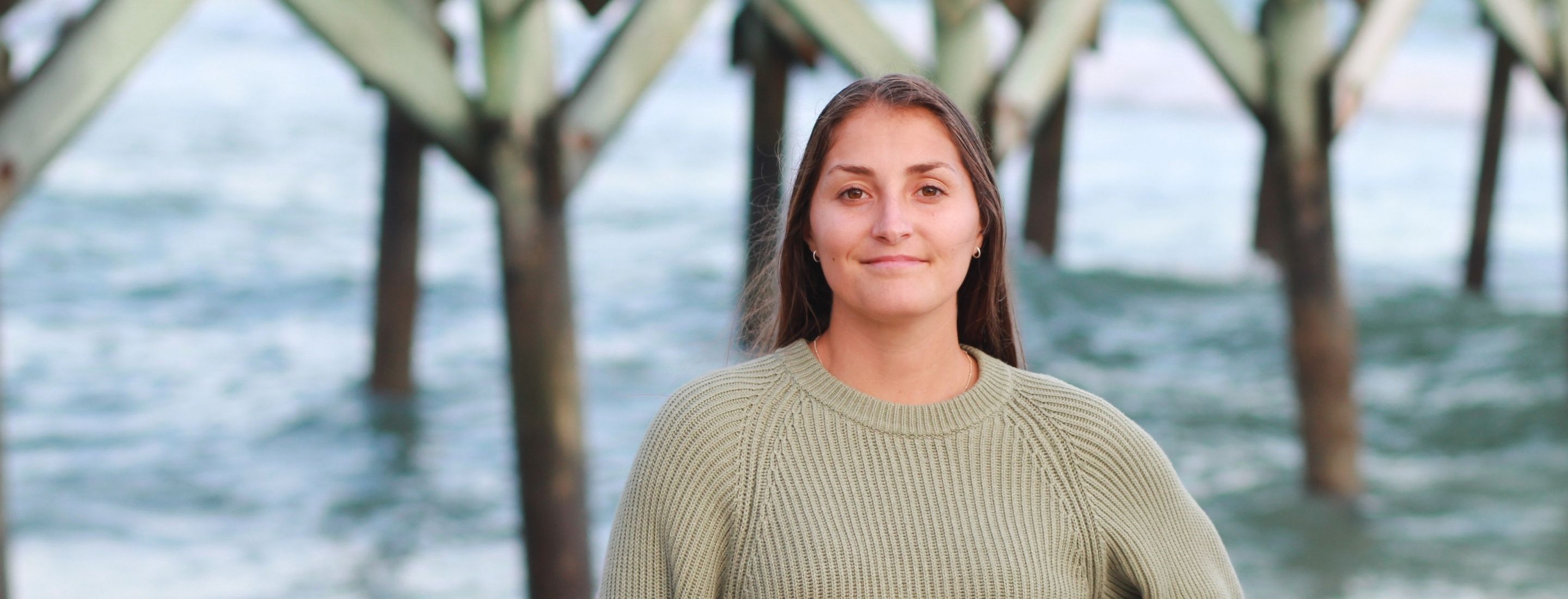 Jessica Cark poses in front of a pier, wearing a beige cardigan.