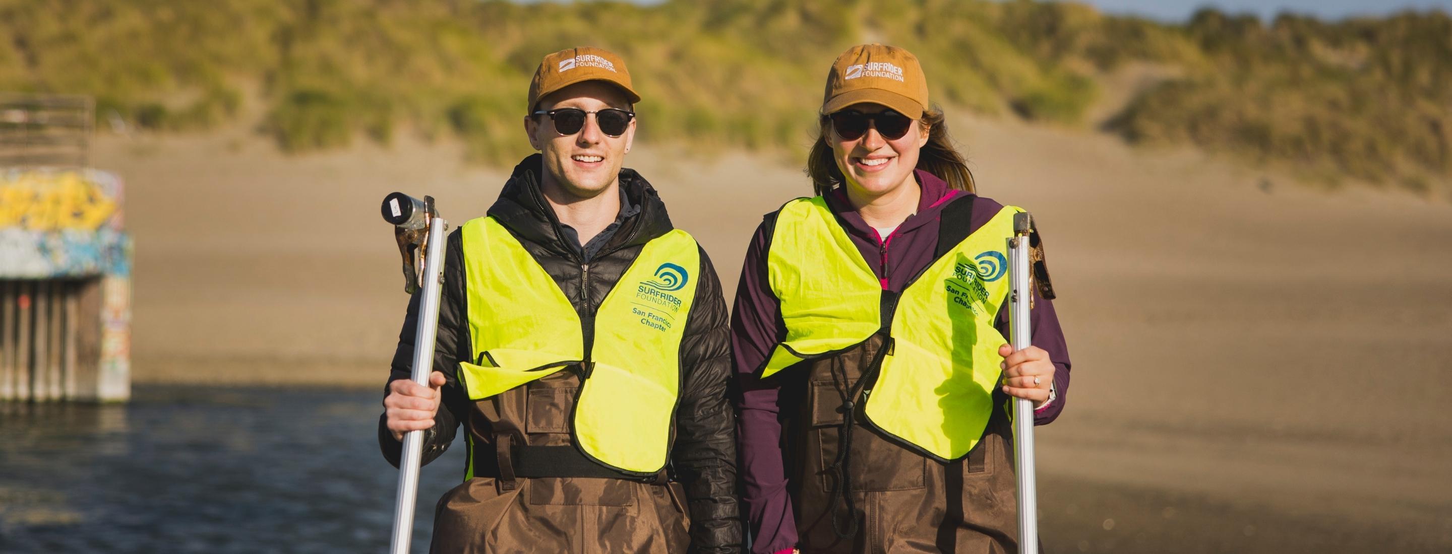 Kyle on left, Amanda on right, pose in hi-viz vests and waders as they prepare to test ocean water quality. They are wearing light brown 