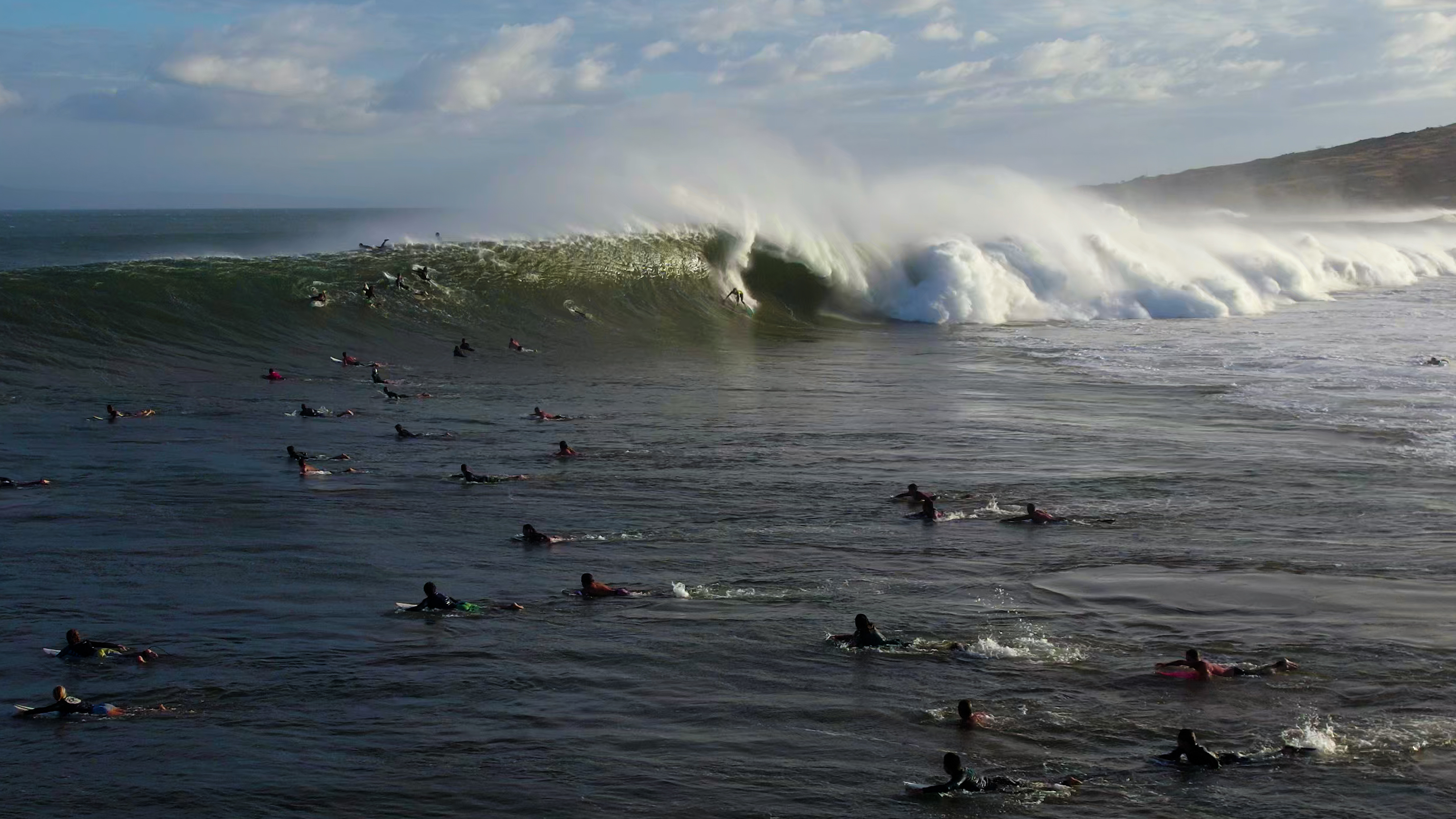 A surfer drops into a fast righthand wave with twenty surfers in the foreground paddling to get out of the way.