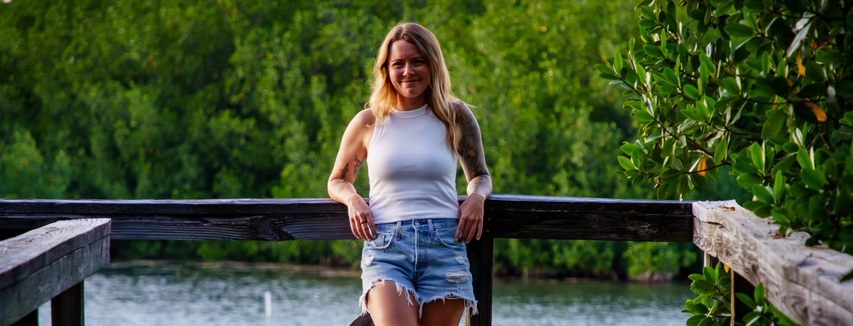 Katie stands on a dock at dusk, the light hitting her right side, she's wearing blue cutoff jeans and a white tank top.