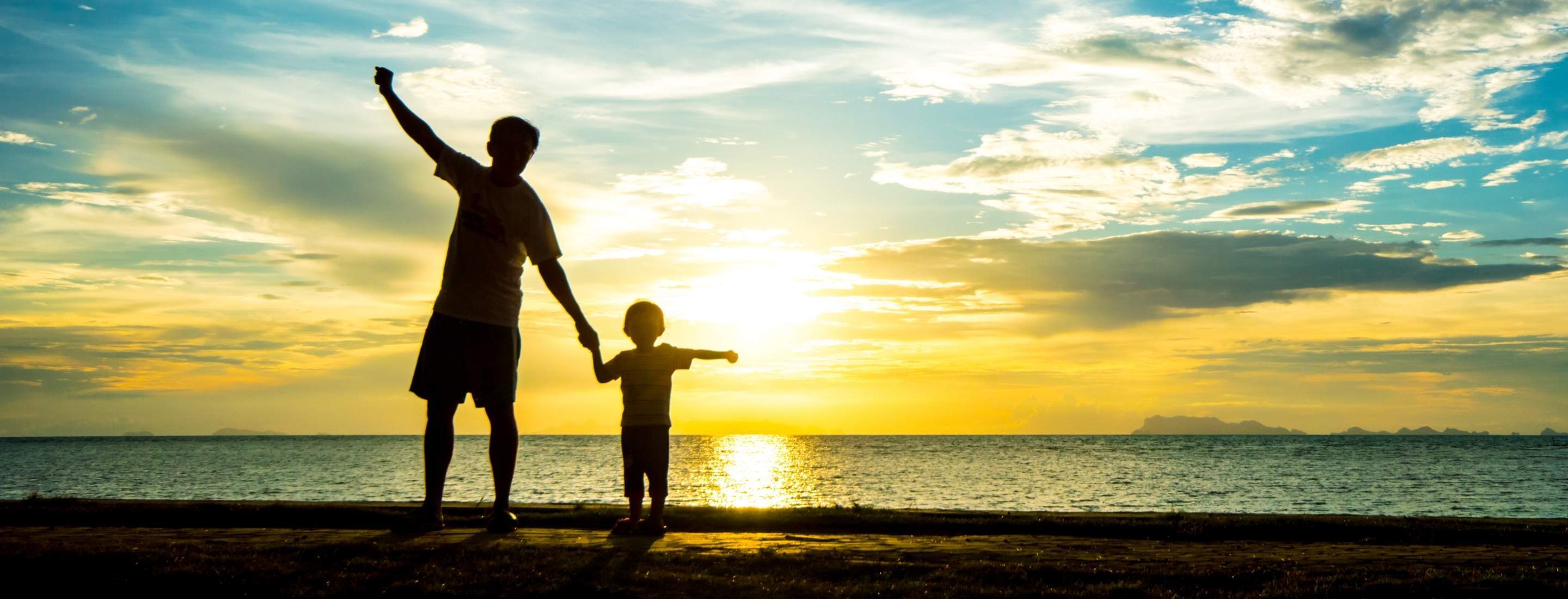 A young and old person hold hands looking on the beach at sunset.